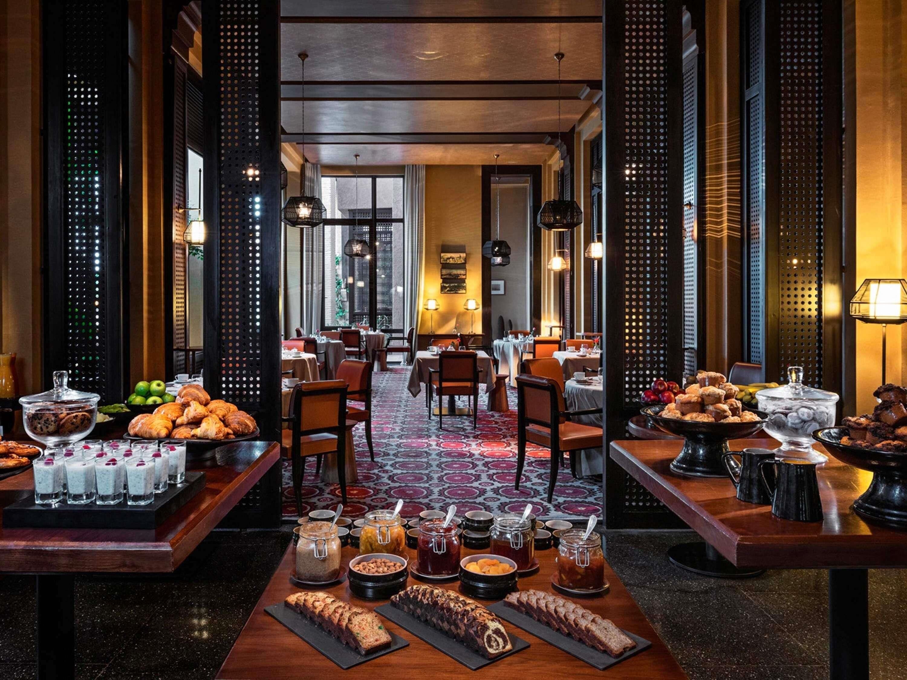 Un comedor de hotel con varias mesas, diversas opciones de comida y bebida, y vistas panorámicas de la ciudad a través de amplios ventanales.