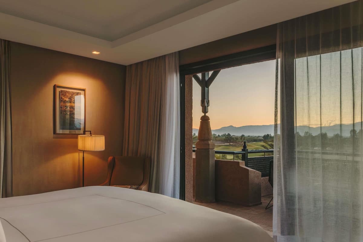 Une chambre d’hôtel dotée d’un grand lit, d’un bureau, d’une chaise, d’une lampe et d’un balcon offrant une vue sur un terrain de golf et les montagnes.