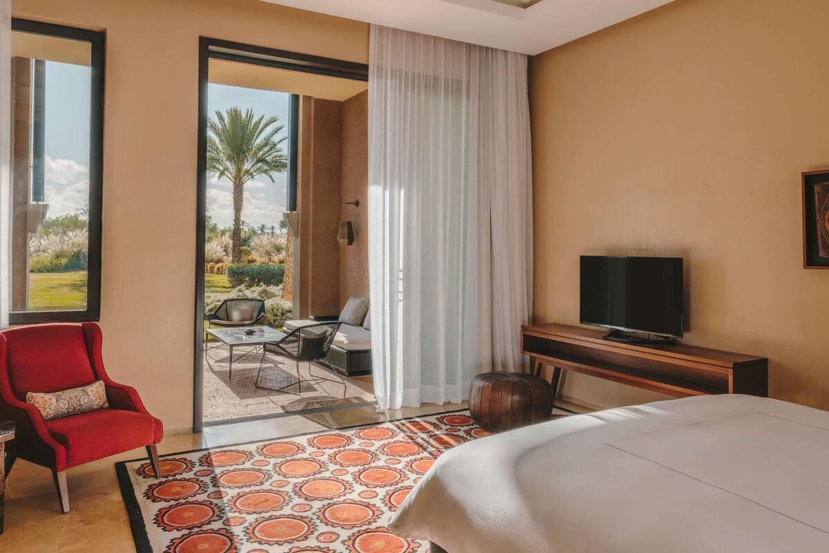 Une chambre d’hôtel avec un lit, une télévision, un fauteuil rouge, un canapé, une petite table et une vue sur un jardin, visible à travers une porte vitrée.