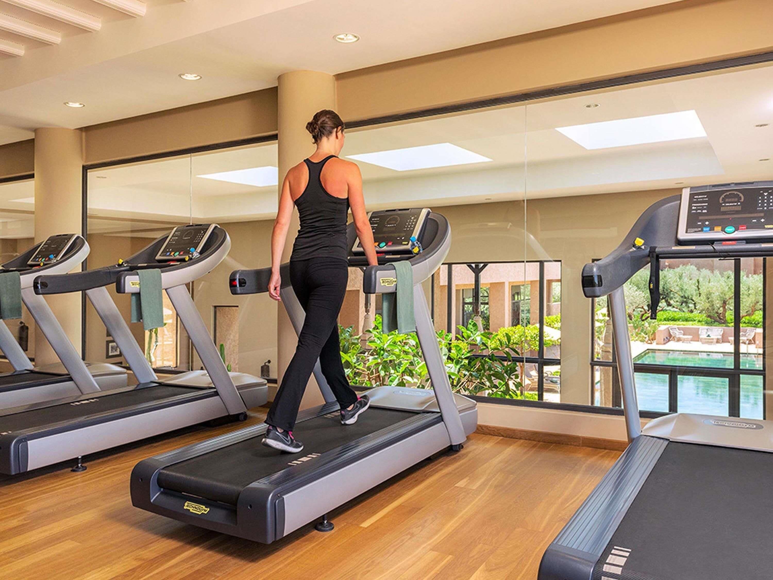 Una mujer camina sobre una cinta de correr en un gimnasio con ventanas amplias y vistas al verdor exterior.