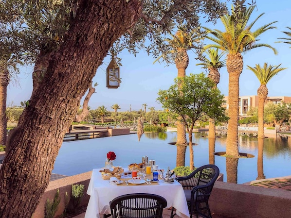 Mesa dispuesta con alimentos para el desayuno, con vista a un cuerpo de agua tranquilo, rodeada de palmeras y un cielo despejado.