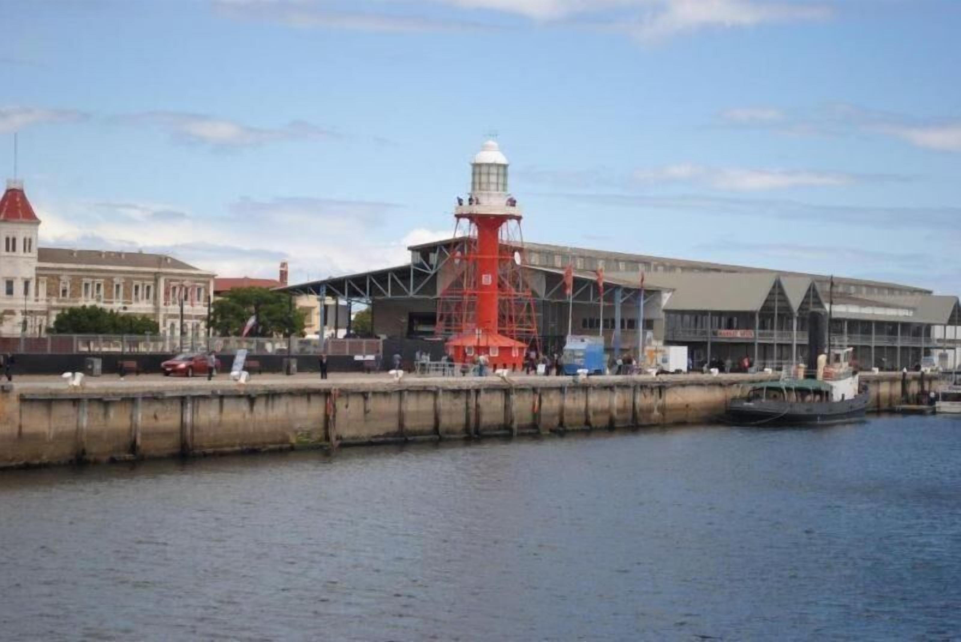 Vista al agua con un faro rojo destacado, un gran edificio industrial y un río tranquilo.