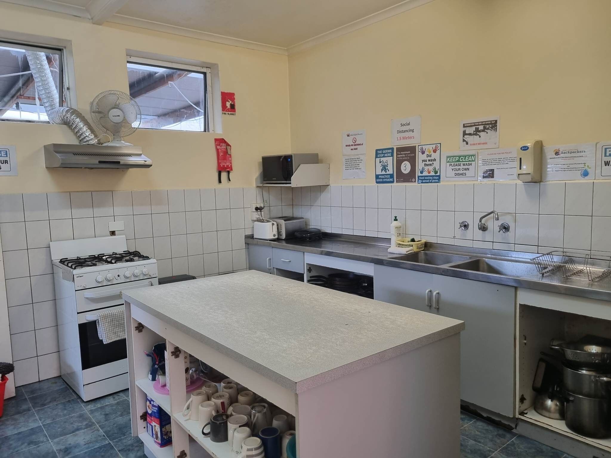A compact kitchen with a gas stove, a microwave, a toaster, and a sink. There are various hygiene posters on the wall, and a small shelf with mugs.