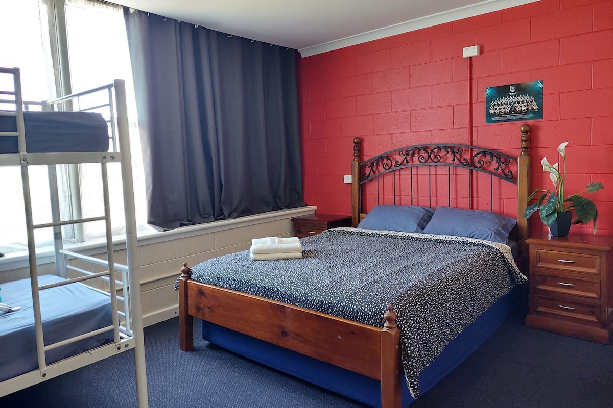Ein Schlafzimmer mit Etagenbett, einem Einzelbett mit gemustertem Bettbezug, einer roten Backsteinwand, einem Fenster mit Vorhängen und einem kleinen Nachttisch mit Pflanze.