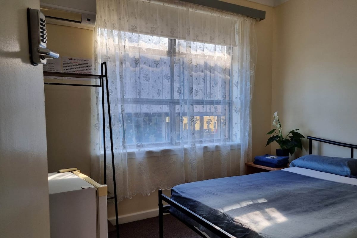 Ein Schlafzimmer mit einem Bett, einem Schreibtisch, einem Stuhl, einer Zimmerpflanze und einem Fenster mit Vorhang.