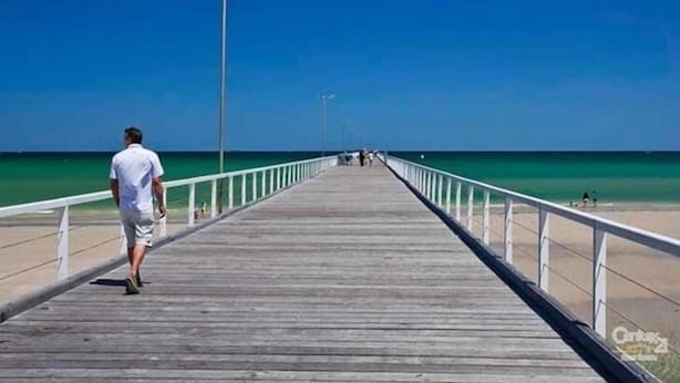 Un muelle de madera se adentra en un mar azul y transparente, con una barandilla a un lado y una persona alejándose de la cámara.