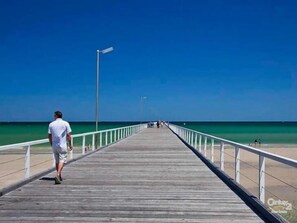 Un muelle de madera se adentra en un mar azul y transparente, con una barandilla a un lado y una persona alejándose de la cámara.
