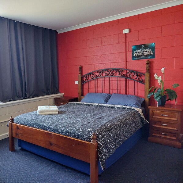 A single-bed room with a red brick wall, a wooden headboard, a blue bedspread, and a nightstand with a plant.