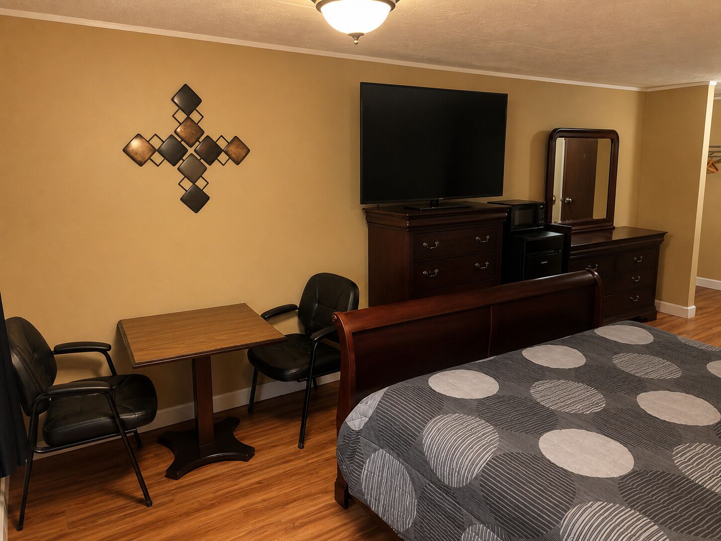 A hotel room with a bed, a desk, two chairs, a dresser, a television, and a wall-mounted decorative piece.