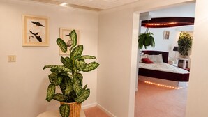 Corredor que dá para um quarto com cama, planta em vaso e obras de arte na parede.