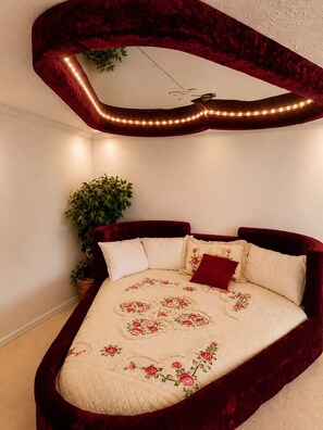 A bed with a floral quilt, a red velvet headboard, and a mirror with lights. - Chateau Royale Inn (Lake Geneva)