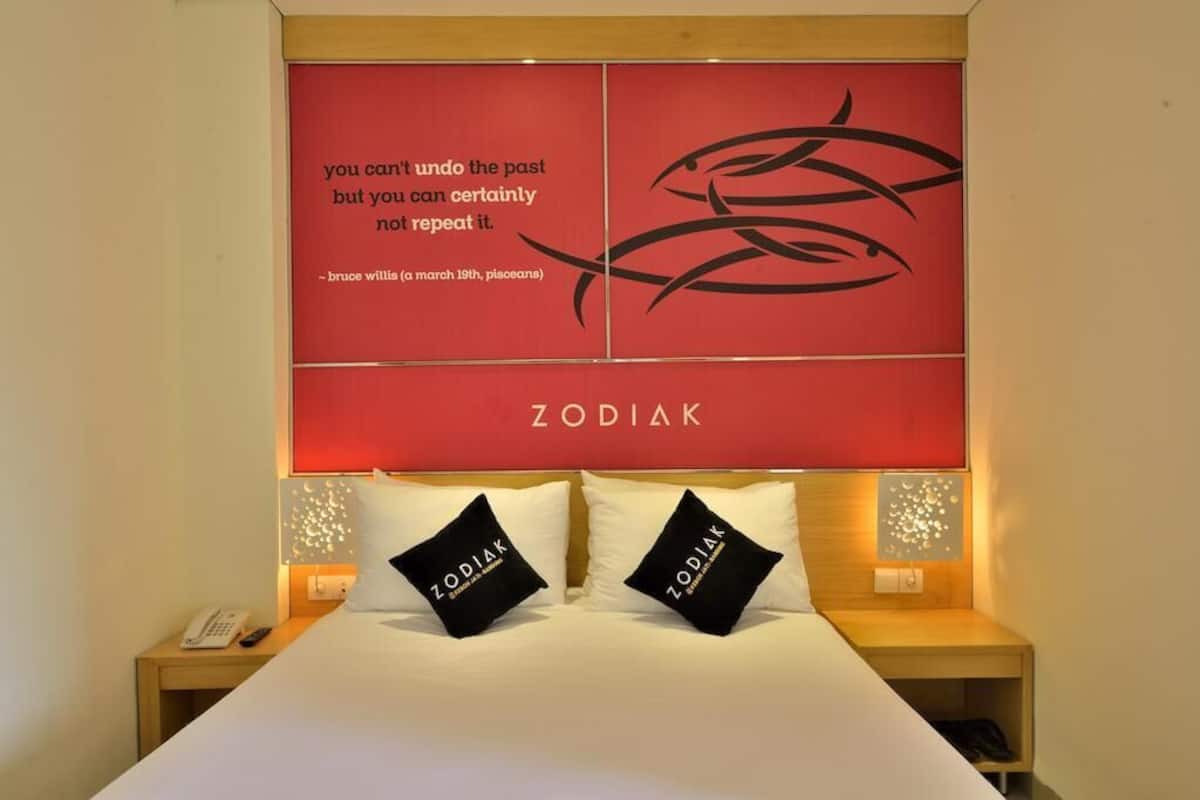 A hotel room with a large bed, two black pillows with 'ZODIAK' text, a red wall with a quote and fish design, and a wooden nightstand with a phone.