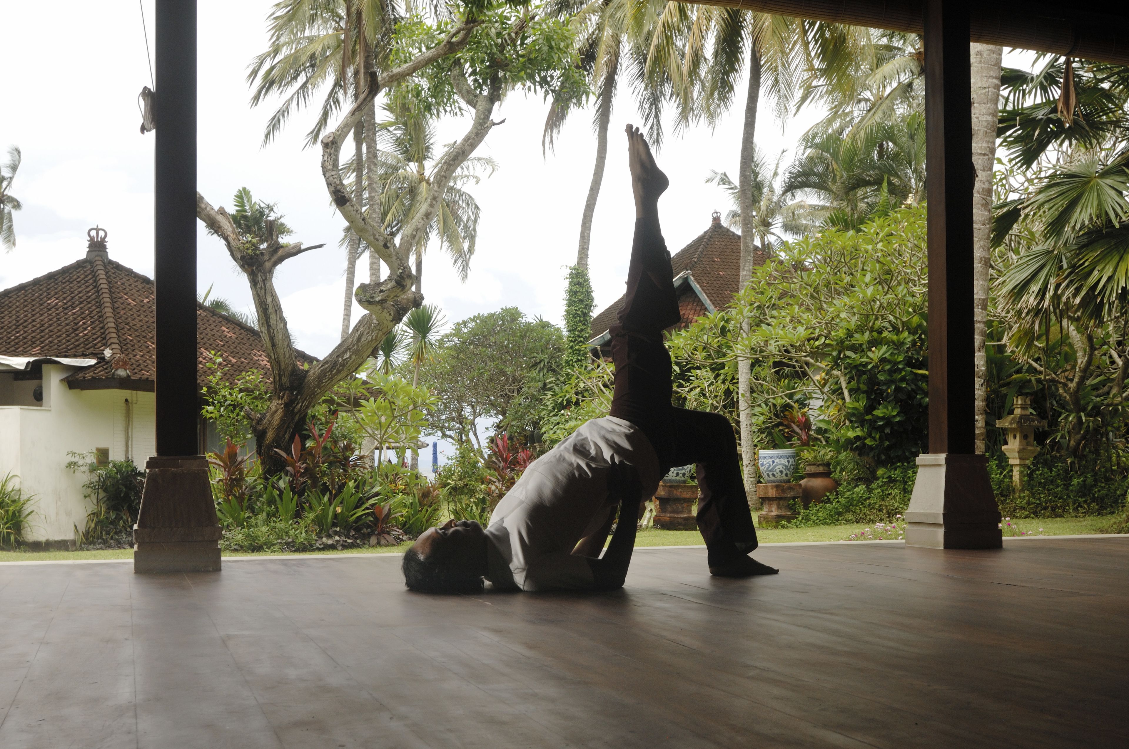 A person performing a yoga pose in a covered outdoor area with a view of palm trees and a building in the background.