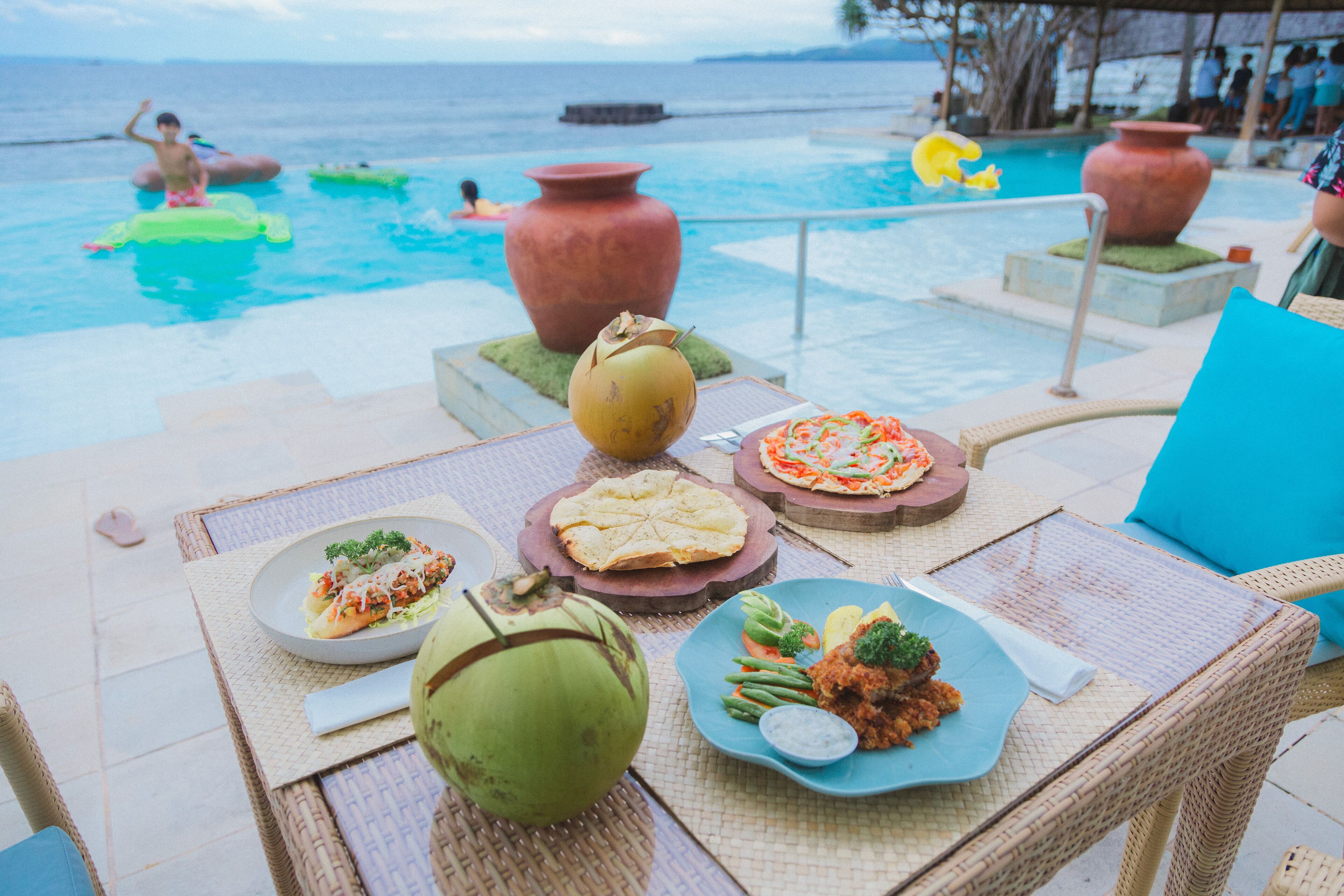Área de comedor junto a la piscina con vista al océano, mostrando una mesa con platos de comida, un coco y jarrones decorativos.