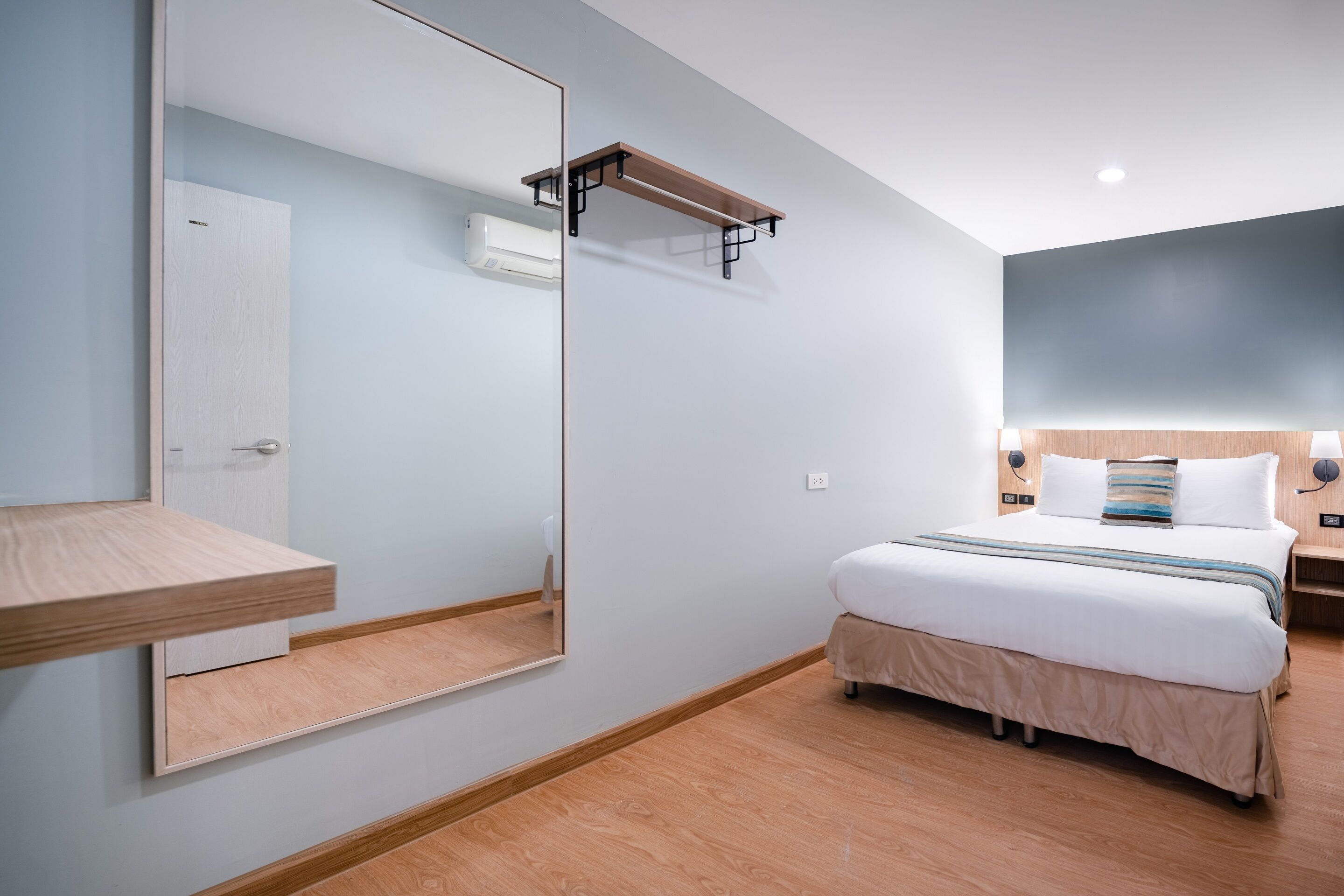 A modern hotel room with a large mirror, a wooden shelf, a bed with white bedding, and a nightstand with a lamp.