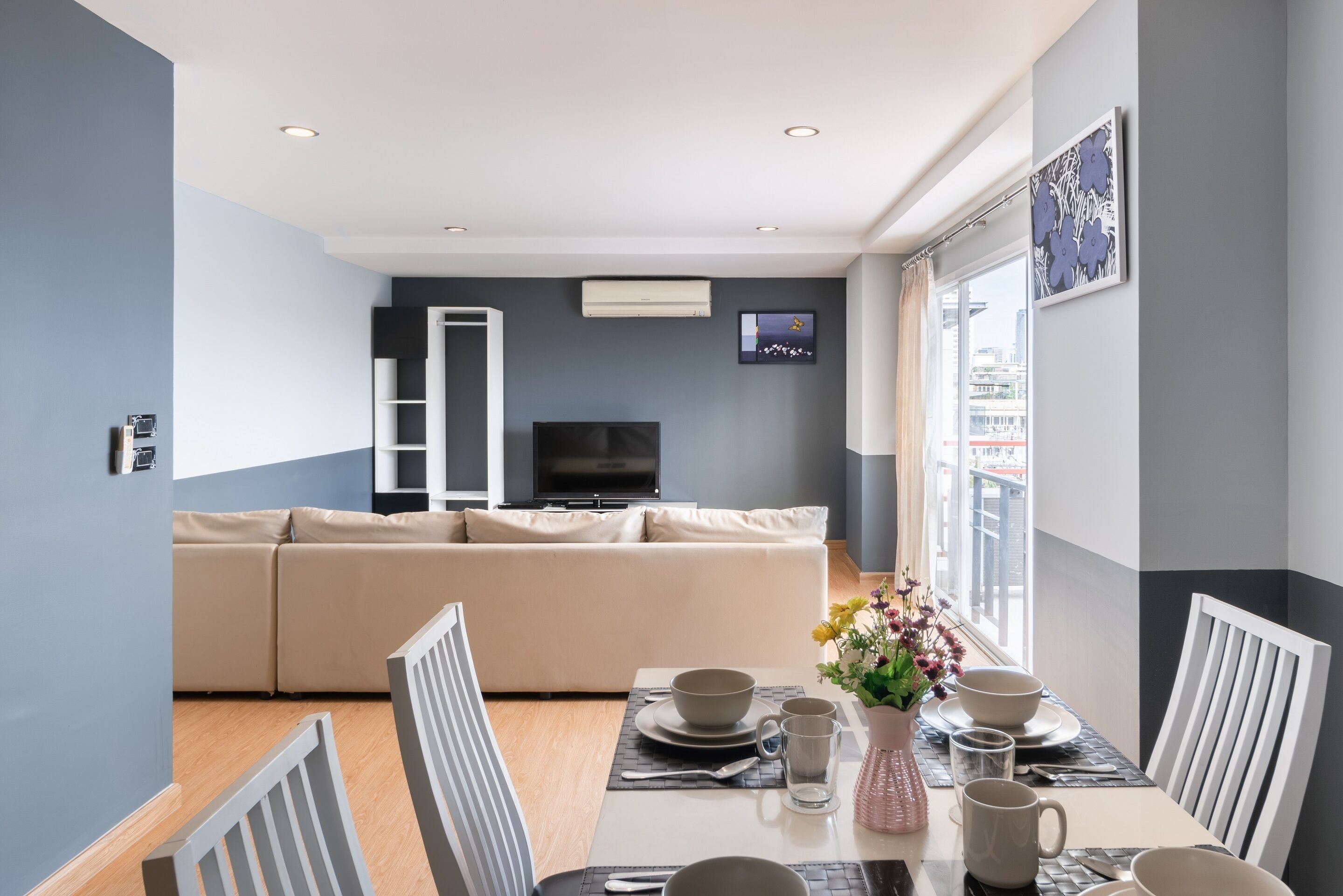 A modern living room with a beige sofa, a dining table set with bowls and cups, and a TV mounted on the wall.