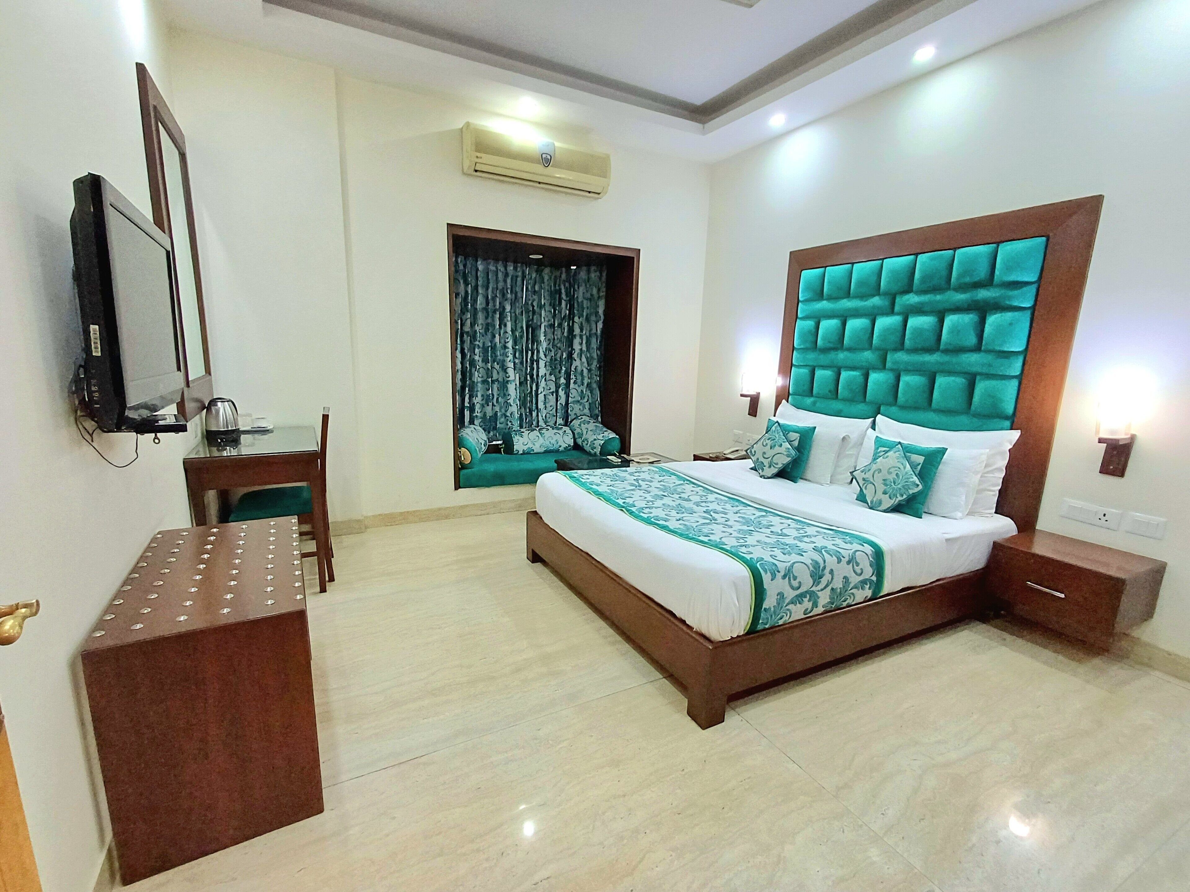 A hotel room with a large bed, a desk, a chair, a TV, and an air conditioning unit.
