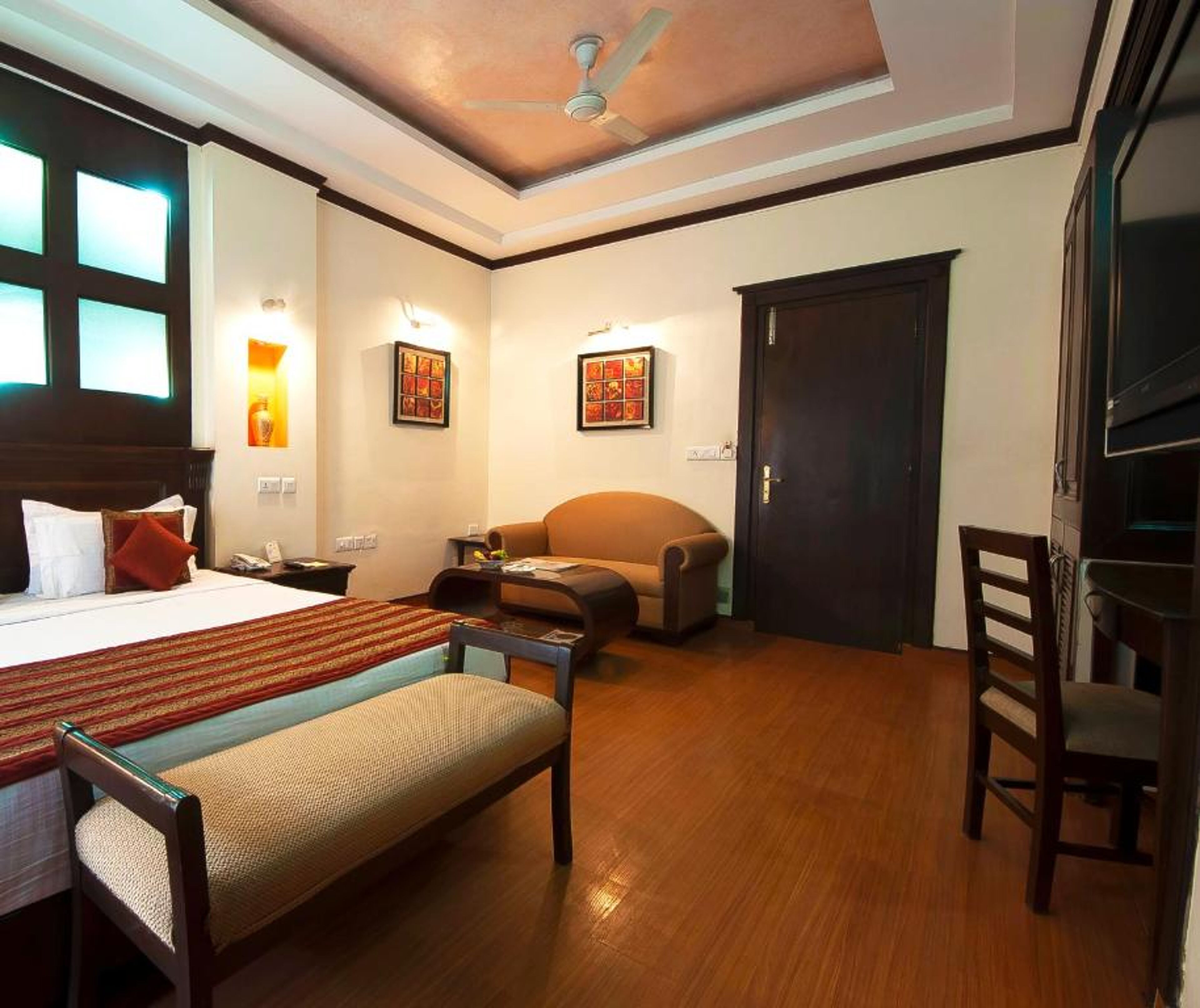 Une chambre d’hôtel avec un lit, un canapé, un bureau et une chaise. Il y a des lampes fixées au mur et un ventilateur de plafond.