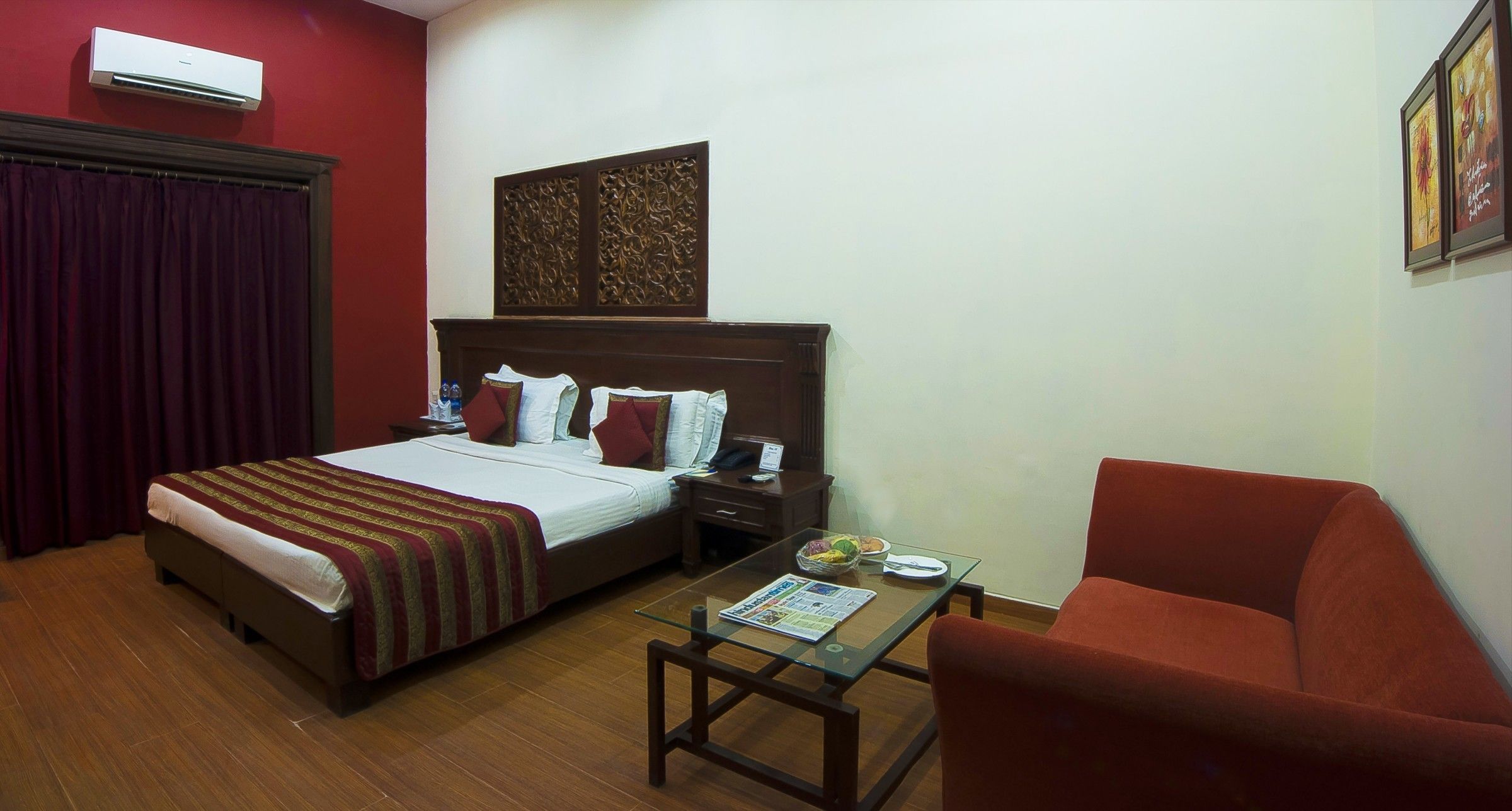 A hotel room with a bed, a glass table with a magazine, a red armchair, and a wooden headboard.