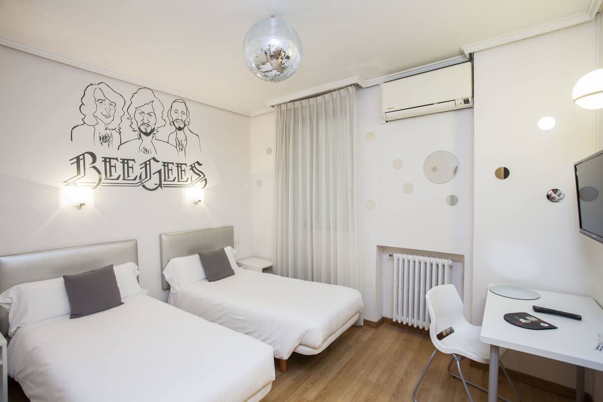Hotelkamer met twee bedden, een bureau met stoel, een televisie en een muur met een mural van de 'Bee Gees'.