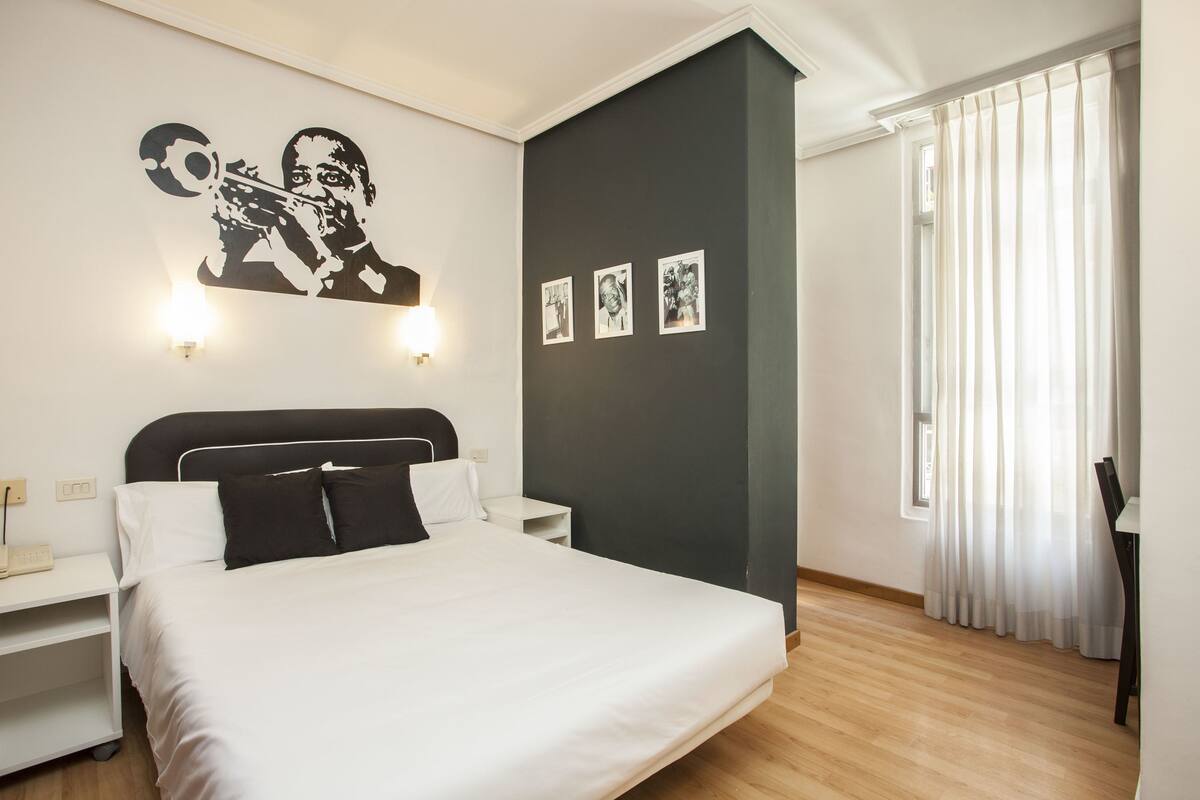 Een keurig opgemaakt slaapkamer met een bed, nachtkastjes, een wandlamp en een raam met dunne gordijnen.