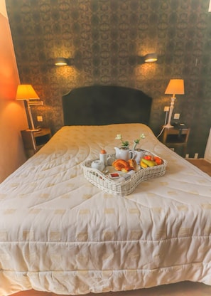 Una cama bien hecha con colcha acolchada, una cesta con productos para el desayuno y dos lámparas de noche a cada lado.