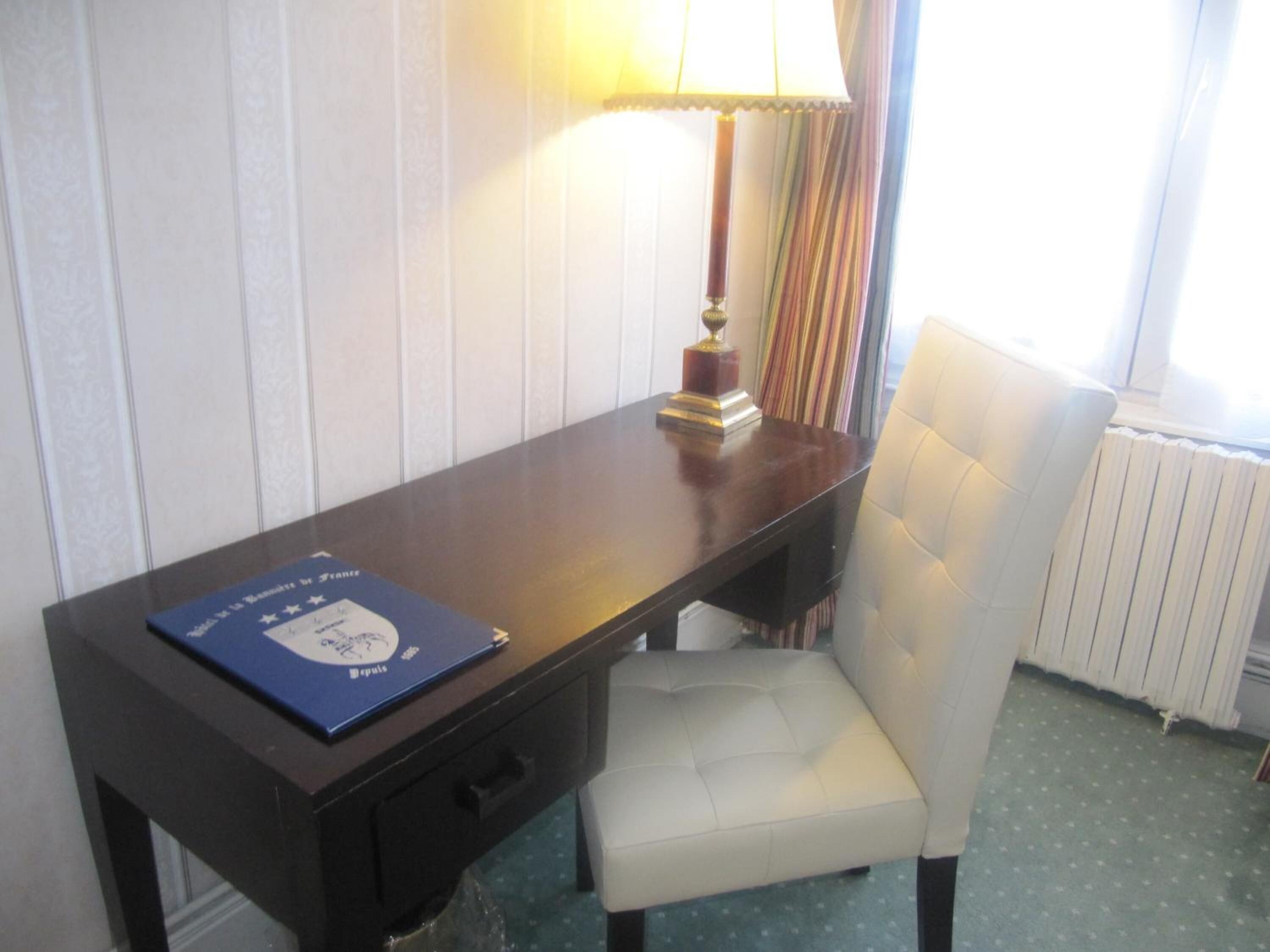 A hotel room with a wooden desk, a white upholstered chair, a lamp, and a blue folder on the desk.