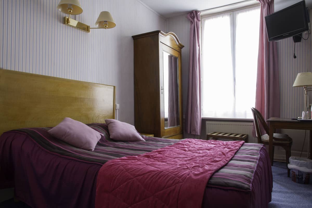 A hotel room with a large bed, a wooden wardrobe, a desk with a chair, a television, and a window with curtains.