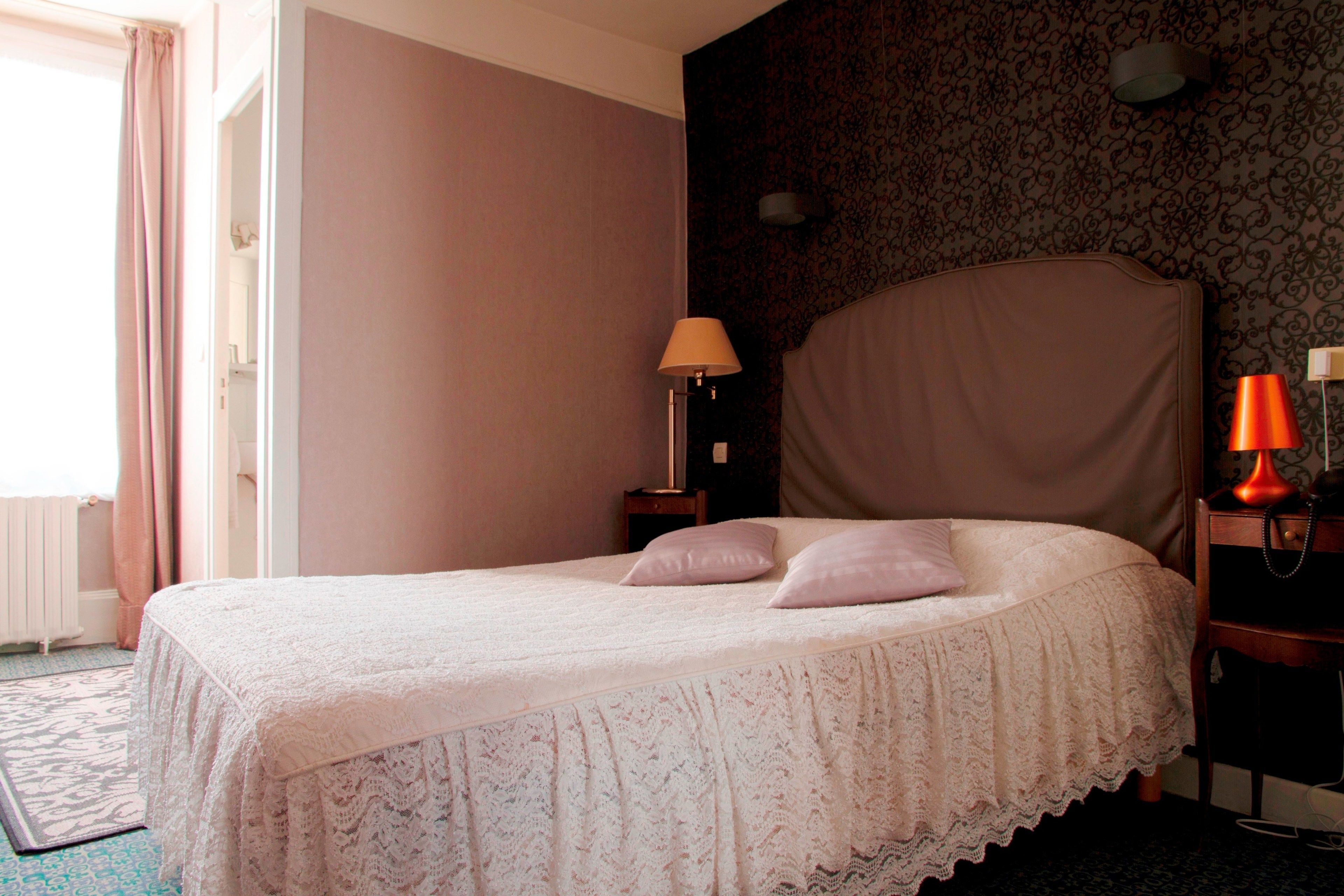 Una cama bien tendida con una colcha de encaje y almohadas, una lámpara de noche y una pared con papel tapiz estampado.