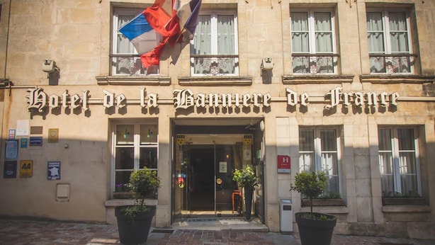 Fachada de hotel con el nombre "Hotel de la Gare de France" bien visible. Hay plantas en macetas en la entrada y banderas sobre ella.