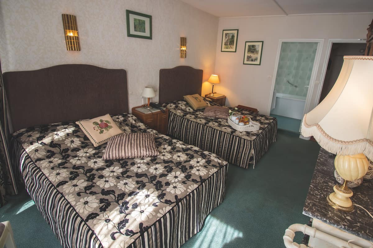 A hotel room with two beds, a nightstand with a lamp, a small table with a lamp, and a bathroom visible through an open door.