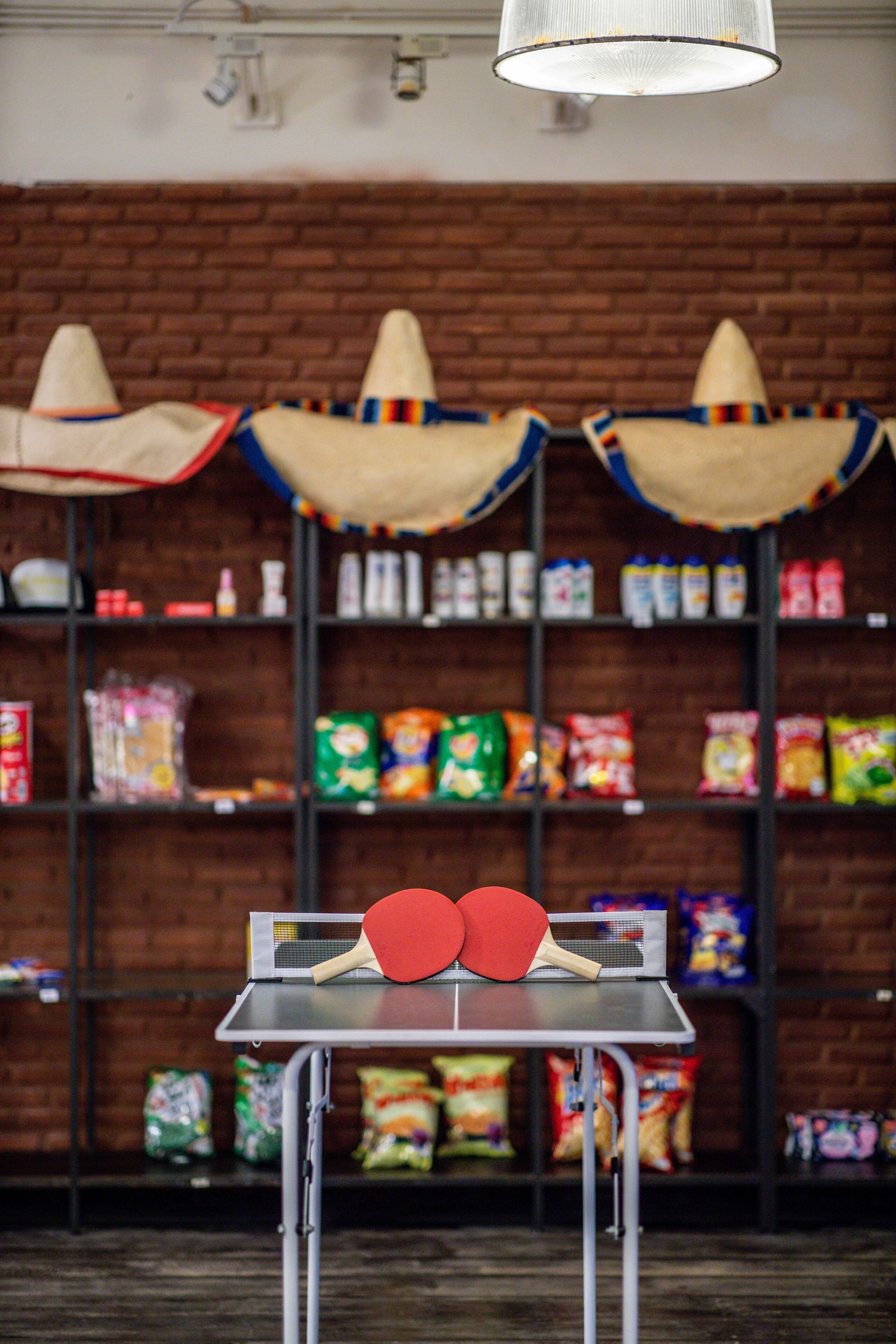 Una mesa de ping pong con dos paletas rojas frente a un estante con aperitivos y alimentos envasados.