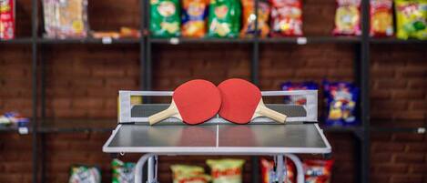 Una mesa de ping pong con dos paletas rojas frente a un estante con aperitivos y alimentos envasados.