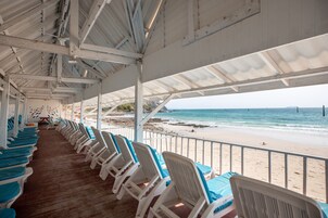 Un área de playa techada con hileras de sillas de plástico blancas y cojines azules, con vista a una playa de arena y el océano.