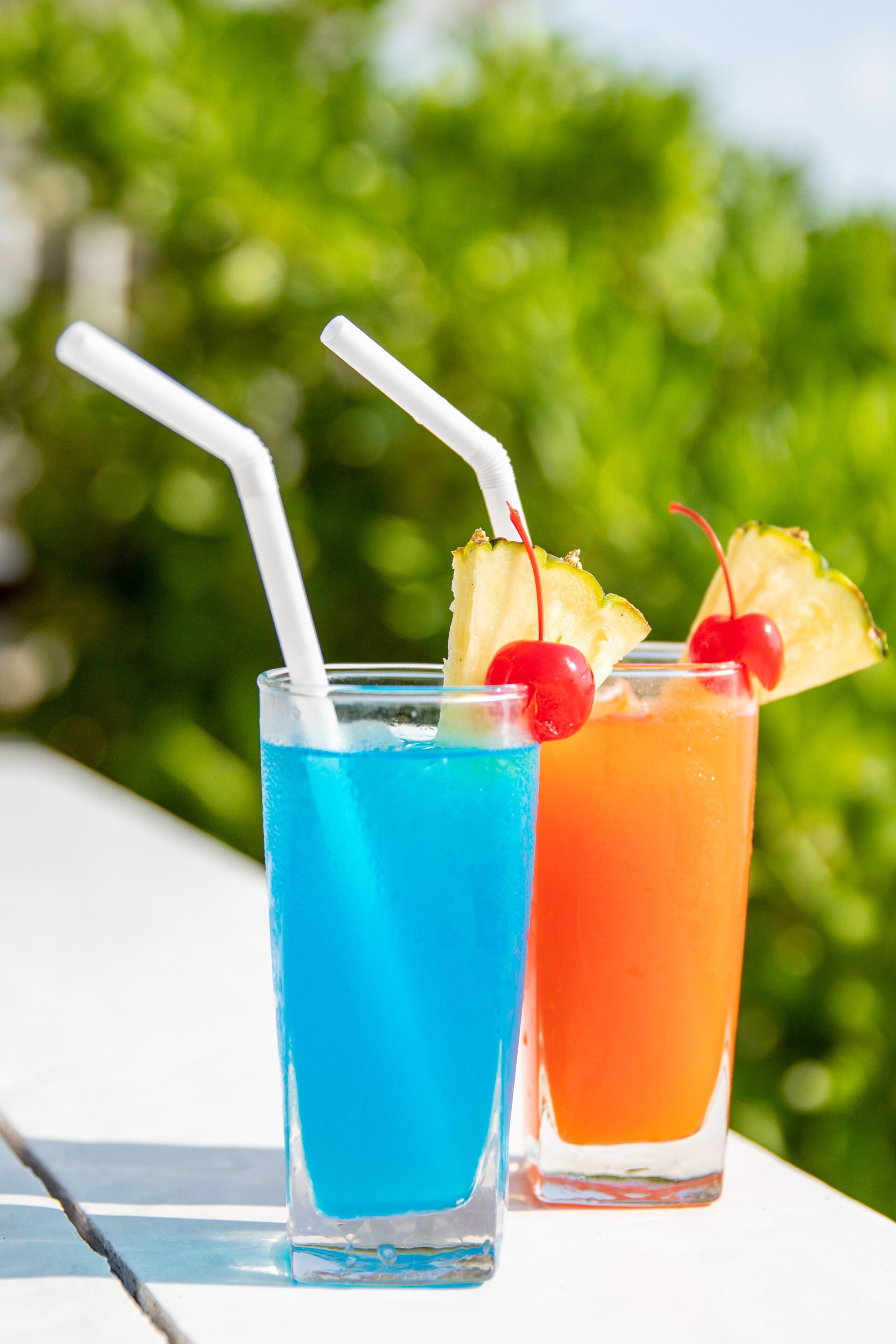 Two glasses of colorful drinks with straws and garnishes, placed outdoors with greenery in the background.