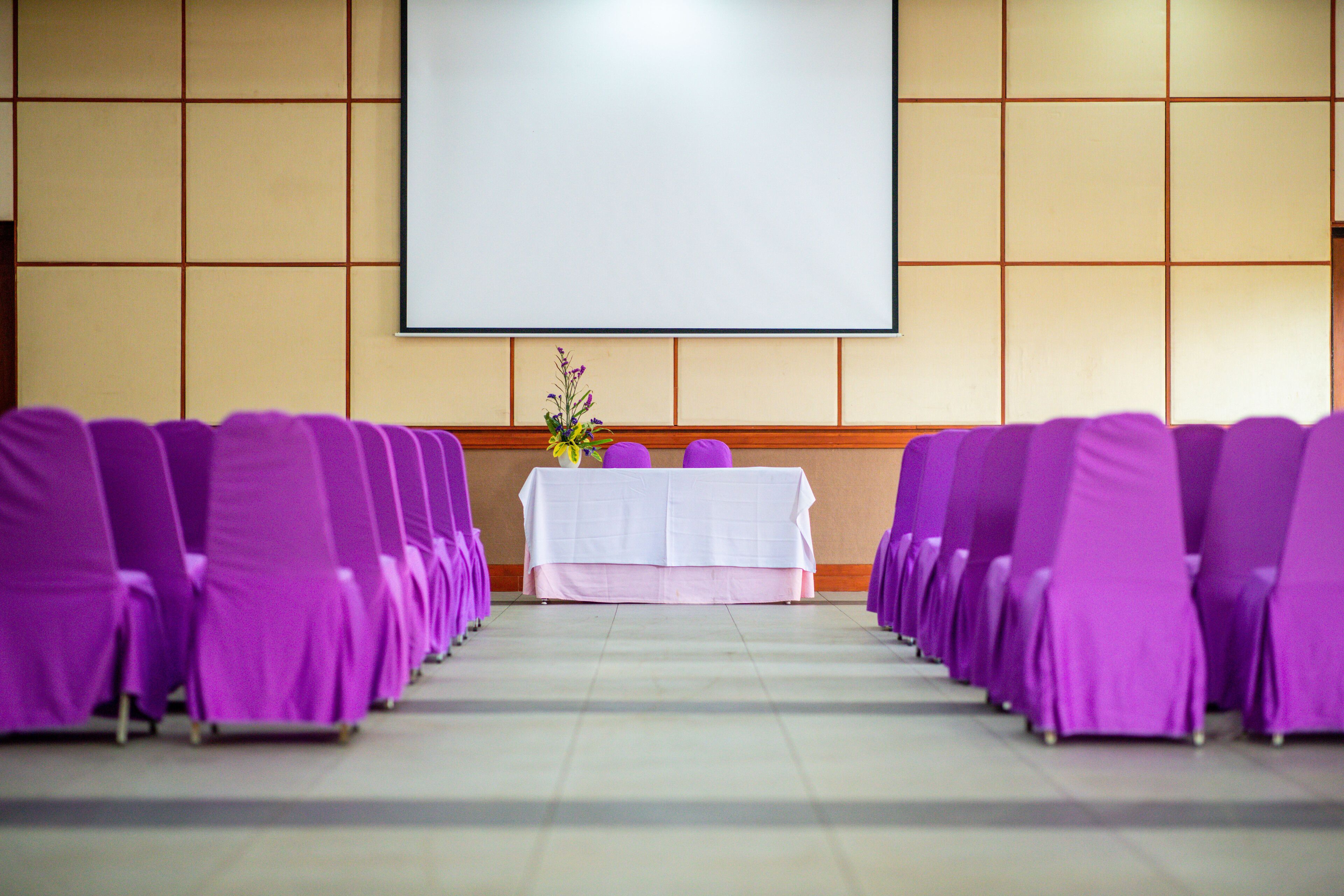 A conference room with rows of purple chairs, a white table with a vase of flowers, and a large blank screen on the wall.