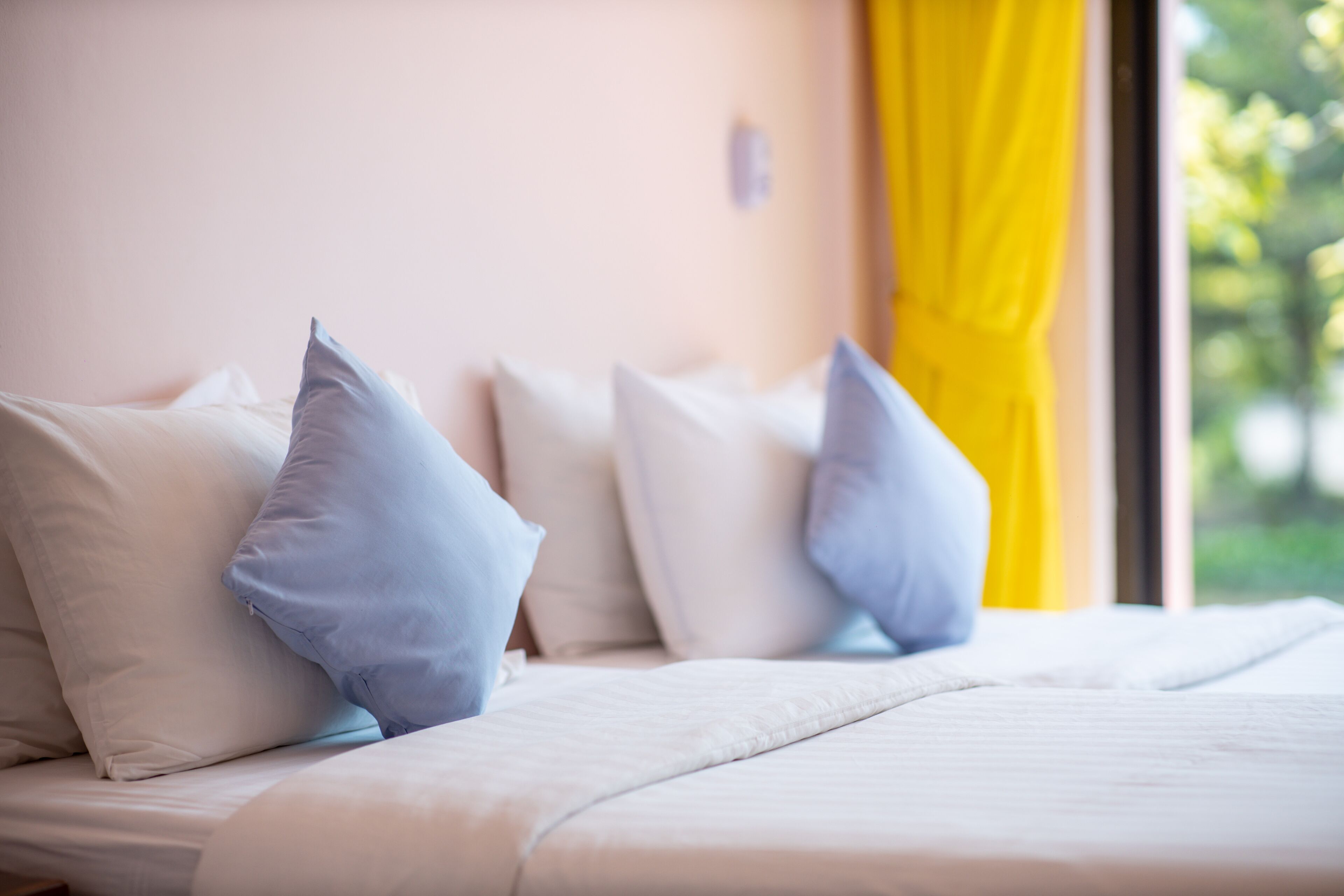 A bed with white bedding and multiple pillows, a window with yellow curtains, and a view of greenery outside.