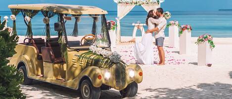 A vintage car parked on a beach with a wedding setup in the background.