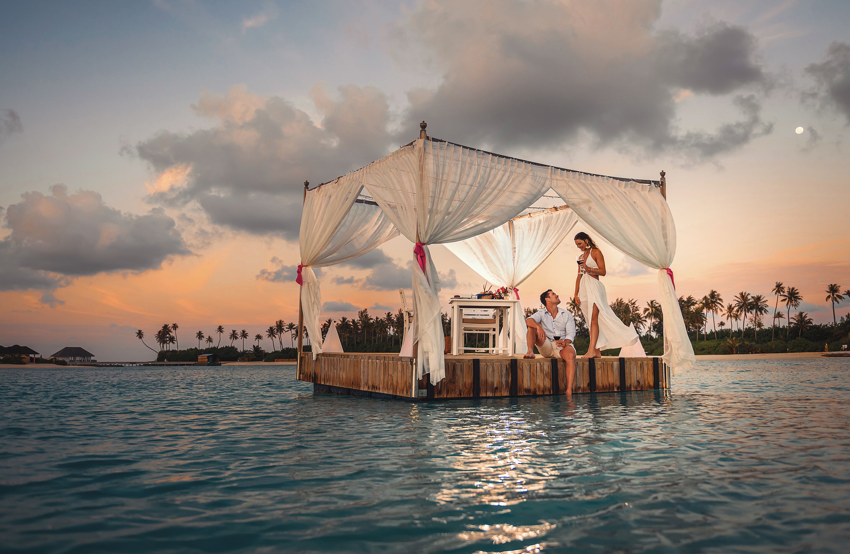 Una plataforma flotante con dosel, rodeada de agua y palmeras, con un atardecer de fondo.