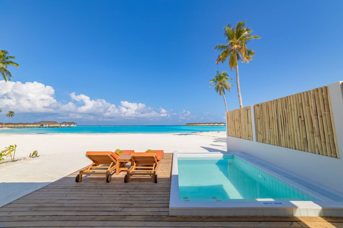 Terraza de madera con sillones de descanso y piscina, con vista a una playa con palmeras y agua azul cristalina.