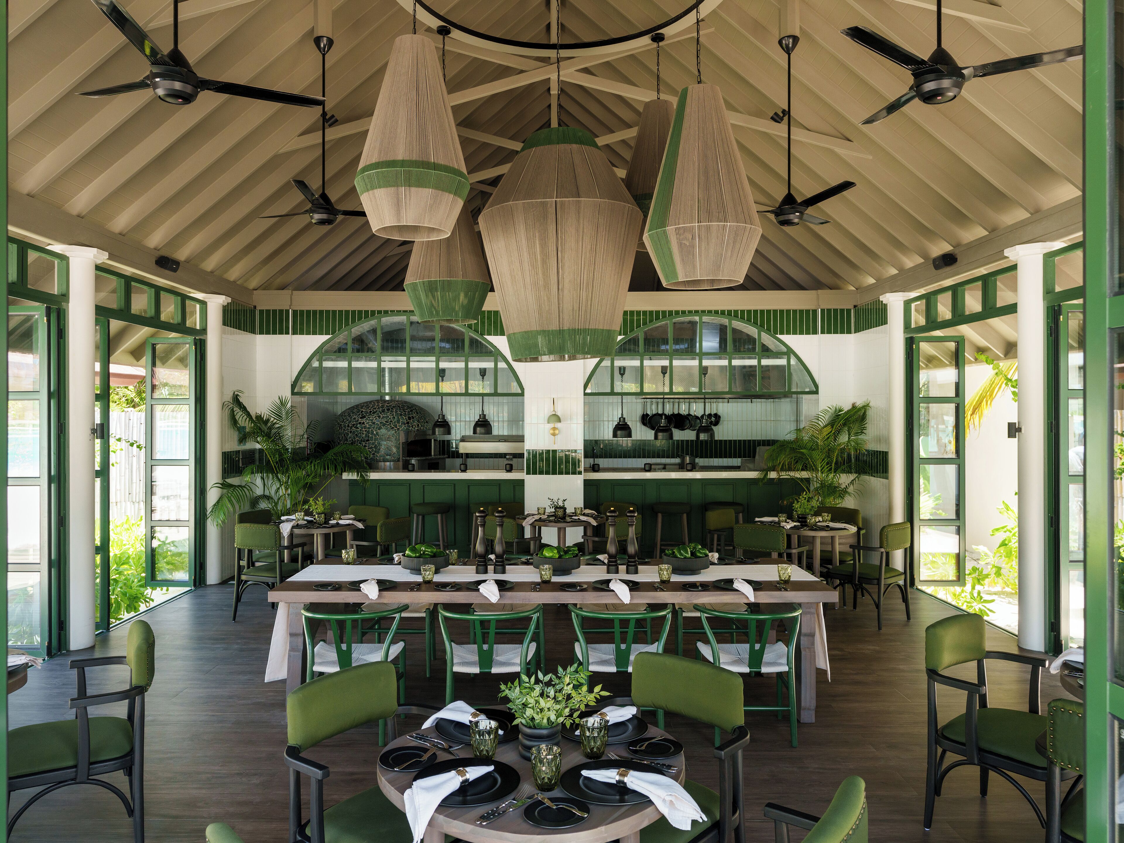 Un restaurant doté de plusieurs tables et chaises, d’un espace cuisine et d’un plafond orné de ventilateurs et de luminaires suspendus.