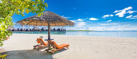 A beach with two sun loungers under a thatched umbrella, a clear blue sky, and a calm sea.