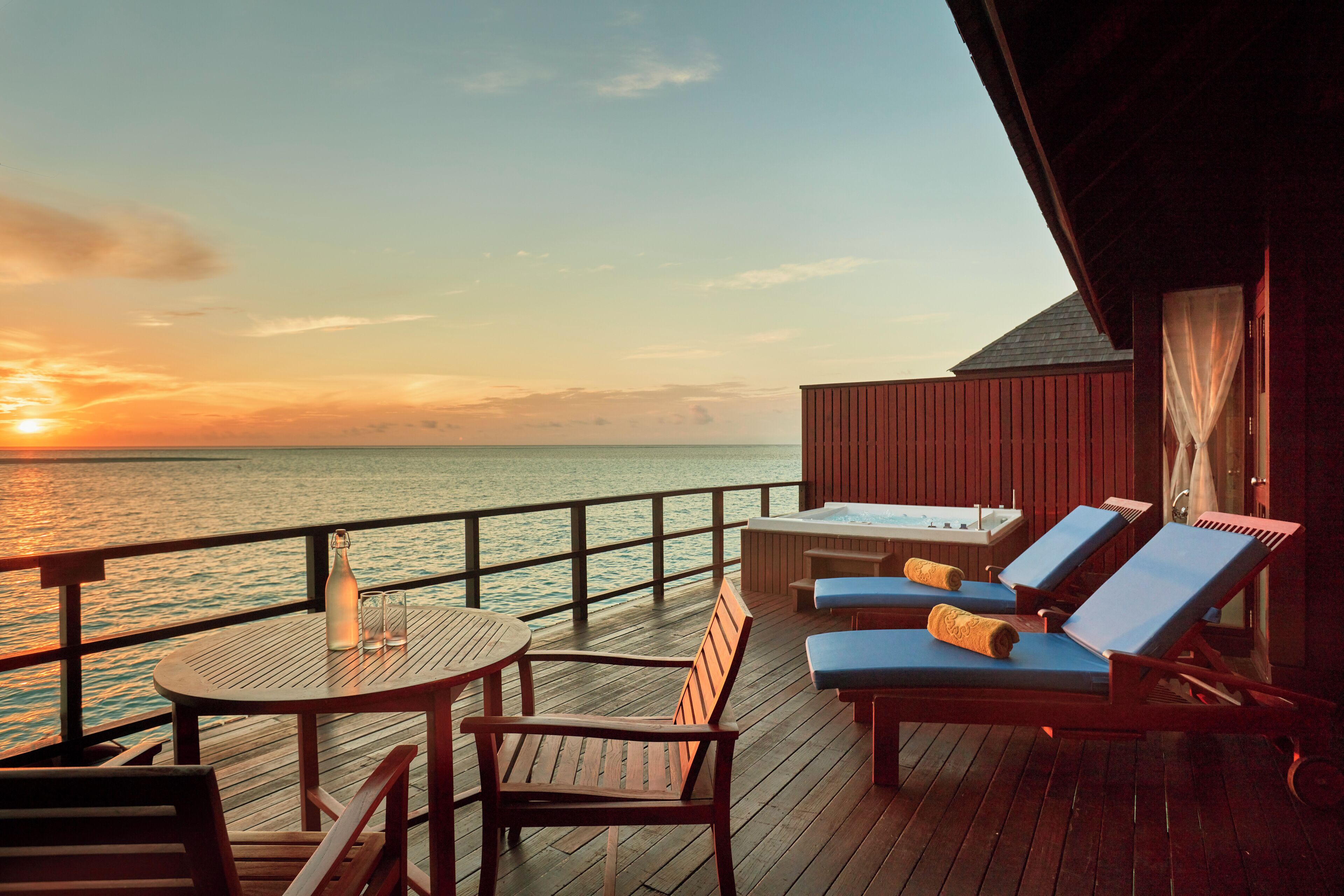 Un pontile di legno con tavolo, sedie e poltrone relax che si affaccia su un corpo d'acqua al tramonto.