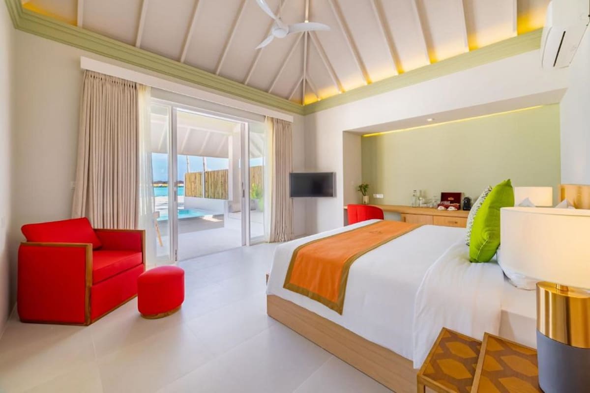 Habitación de hotel con una cama grande, un sofá rojo, un pequeño reposapiés, un televisor, un espejo y un balcón con vistas.