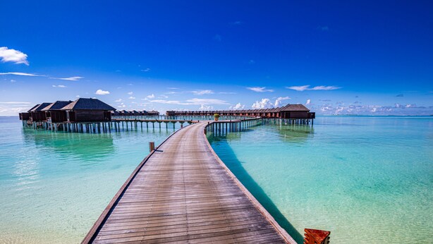 Une jetée en bois menant à des bungalows sur pilotis, au-dessus d’une eau turquoise et cristalline.