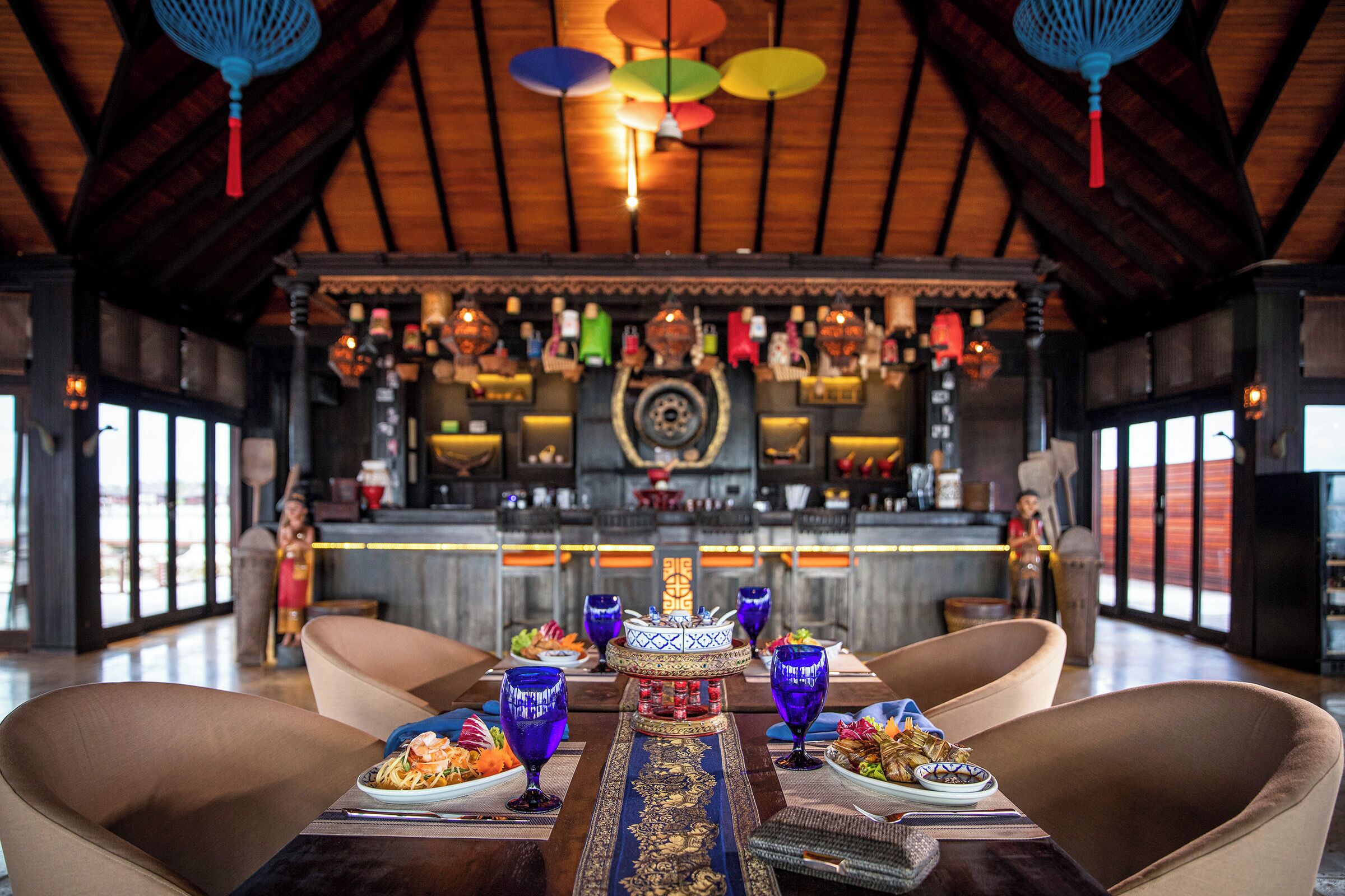 Un restaurante con techo de madera y una zona central de bar. Se observan mesas con copas y platos de comida.