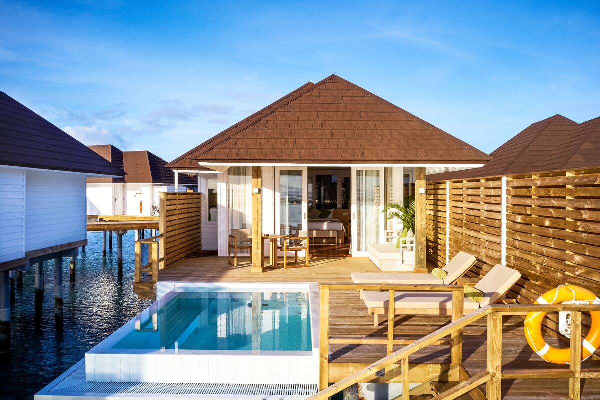 Una cabaña sobre el agua con piscina, terraza de madera y sillones de descanso.