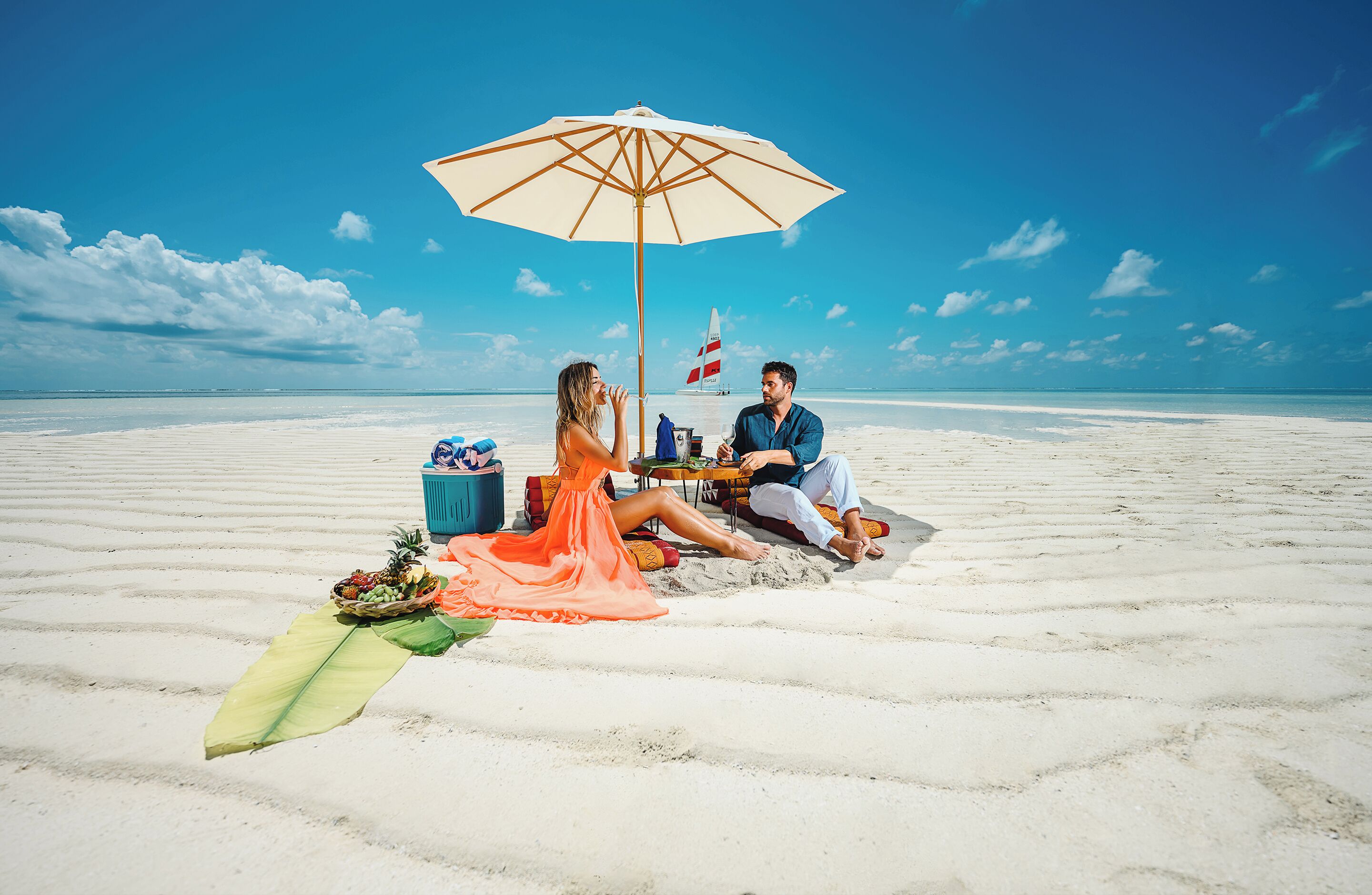 Un couple est assis sur une plage de sable, à l’ombre d’un grand parasol, et on aperçoit un drapeau au loin.