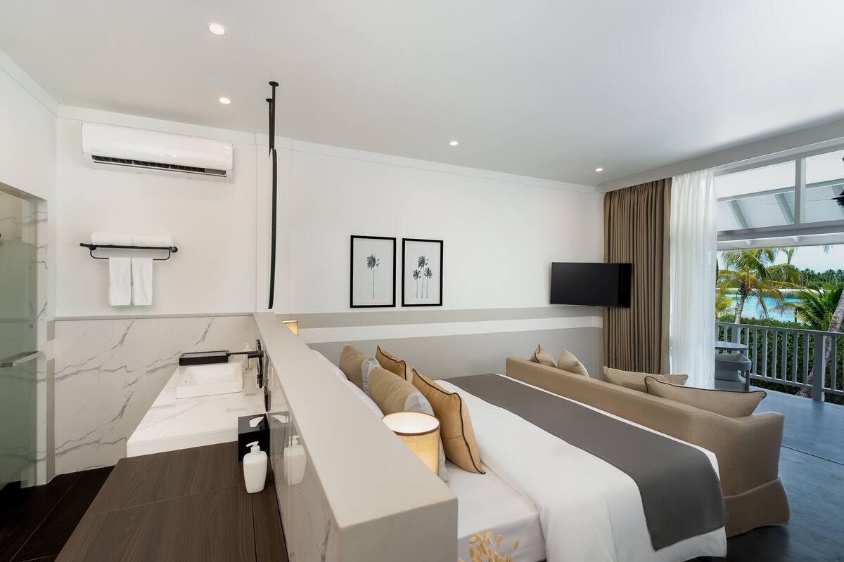 Una habitación de hotel moderna con una cama grande, un sofá, un televisor y un balcón con vistas a palmeras.