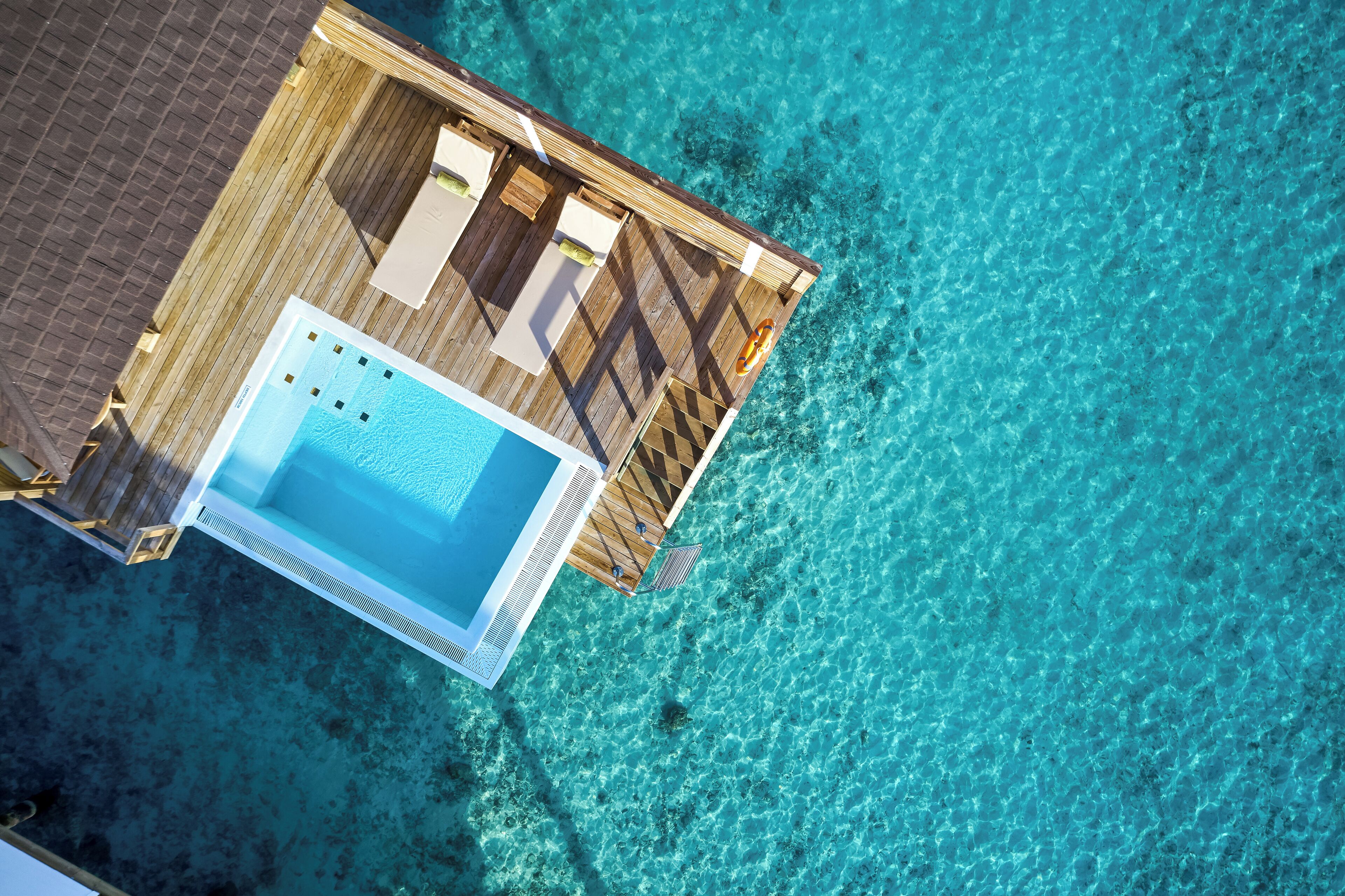 An aerial view of a wooden deck with a pool and lounge area, surrounded by clear turquoise water.